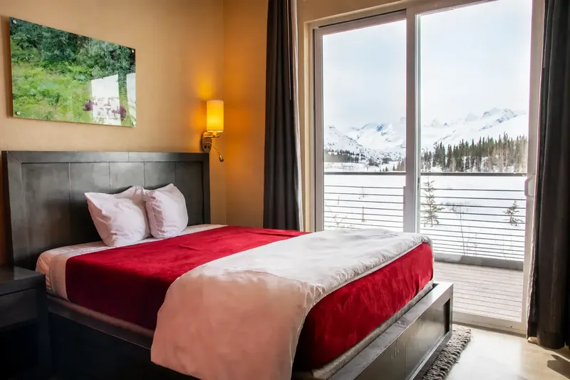A single room at Tsaina Lodge with a Tempur-Pedic queen bed, a bedside reading lamp and a sliding glass door opening onto a private balcony with a view of the snow-covered Chugach Mountains